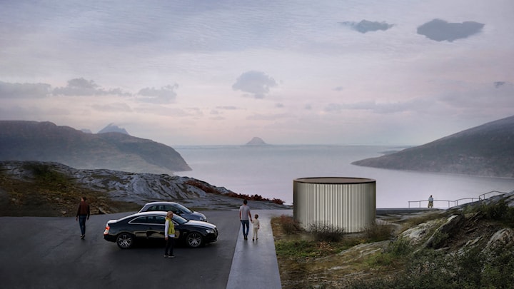 Rundt toalettbygg ved rasteplass med utsikt over fjorden. Noen personer vandrer ved parkeringsplassen der noen bilder står parkert. 