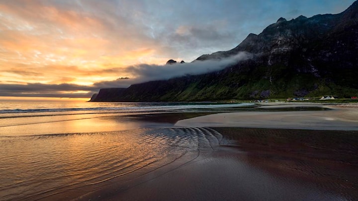 Sandstrand i solnedgang omgitt av hav og spisse fjell.