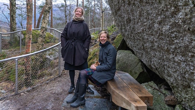 To kvinner sitter på en rastebenk i tre foran en stor stein.